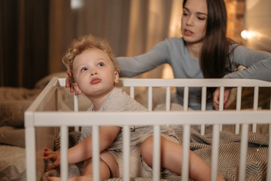 A toddler in a crib with mom sitting on the bed