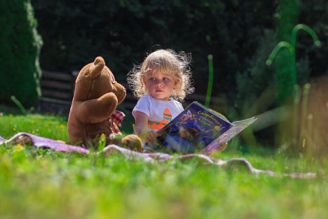 A child reading a book on the grass with a stuffed animal beside
