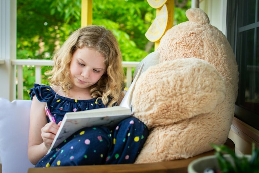 A girl writing in a dream diary beside a big teddy bear.